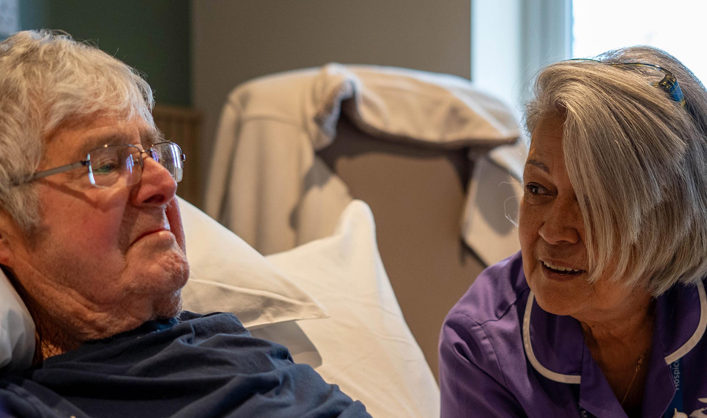 Elderly man in bed holding hands with a nurse in a purple uniform who speaks to him.