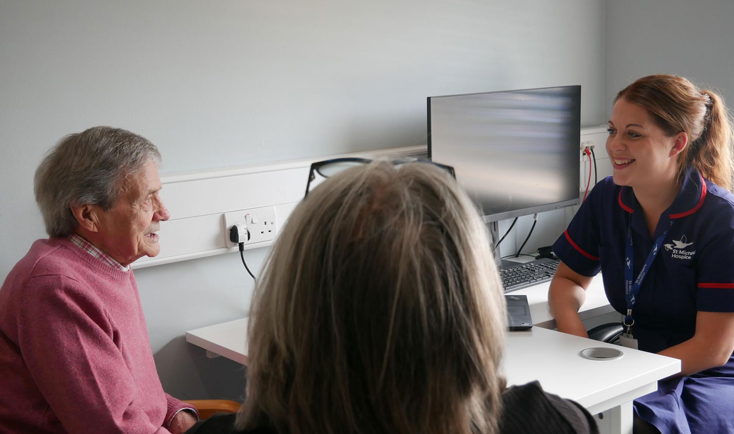 Smiling nurse in blue uniform consults with an elderly patient at a desk.