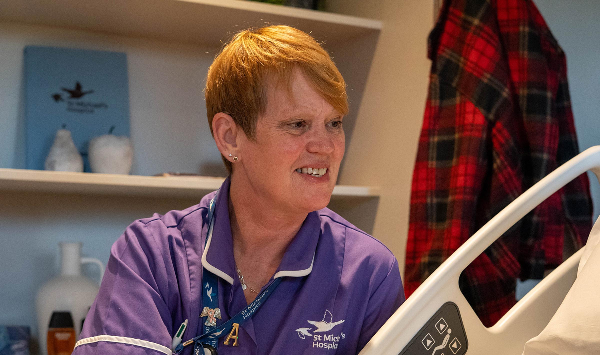 Friendly nurse in purple scrubs smiling next to a hospital bed.