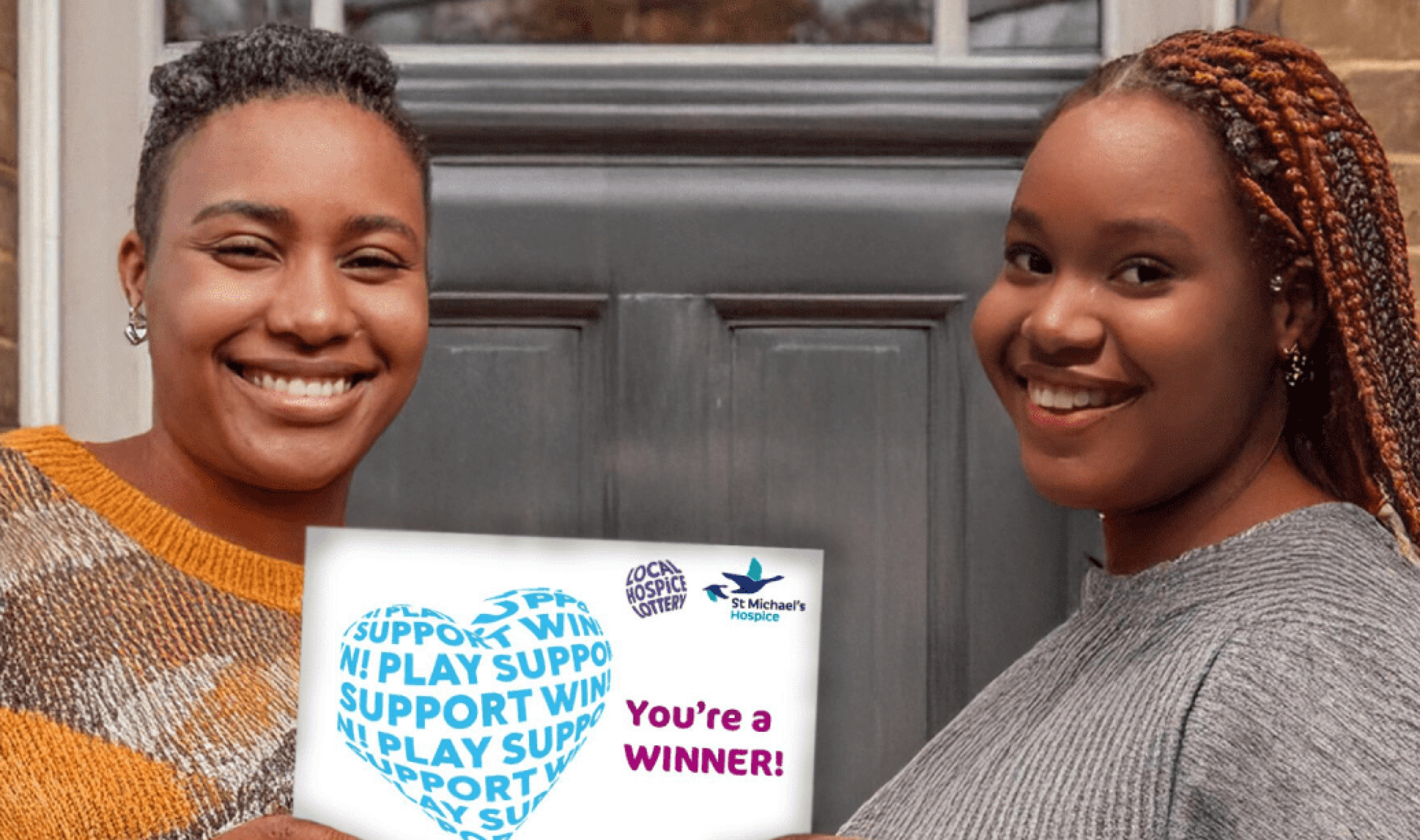 Two smiling women hold a card displaying 'YOU'RE A WINNER' outside a brick house.