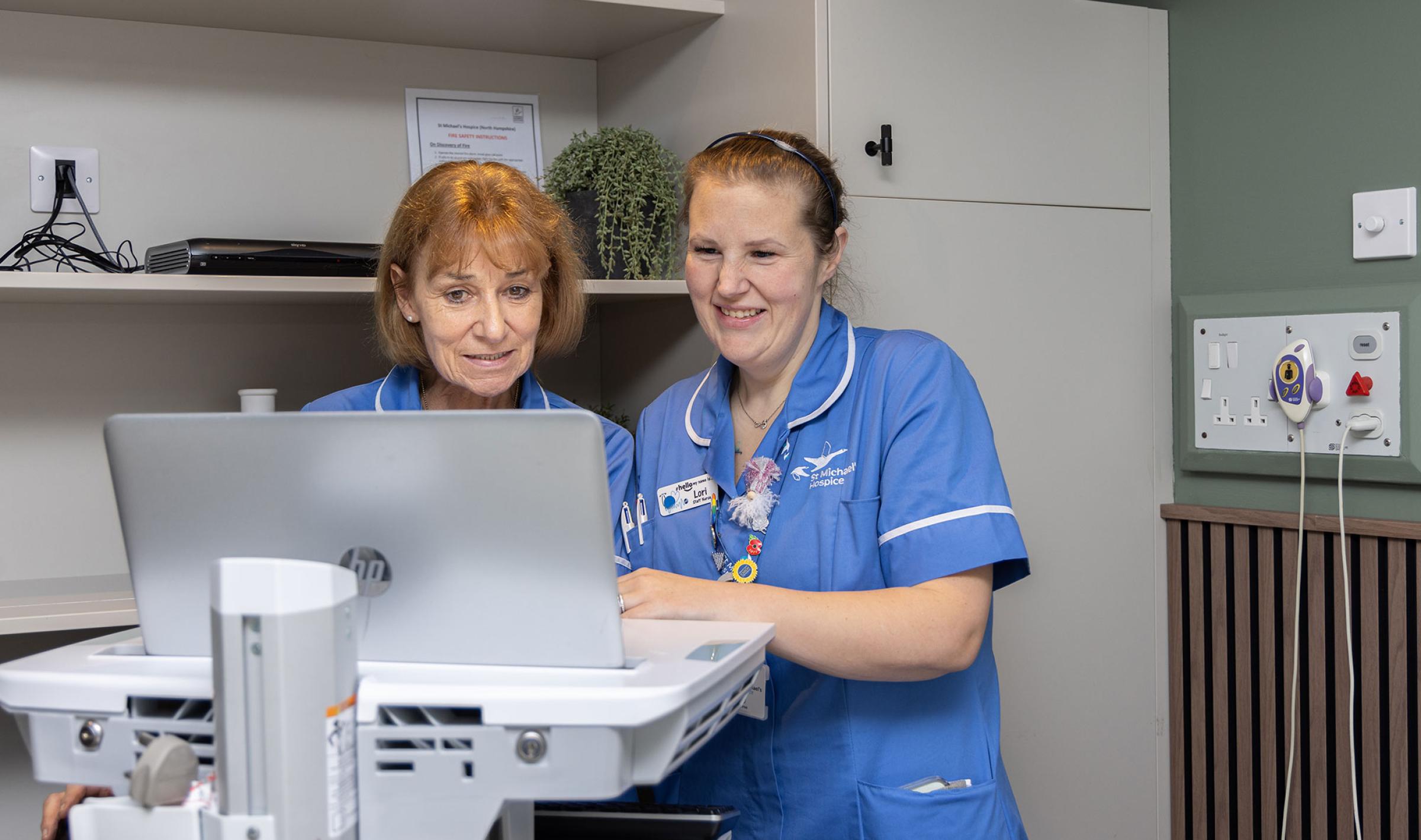 Two smiling nurses viewing a laptop in a clinical setting.
