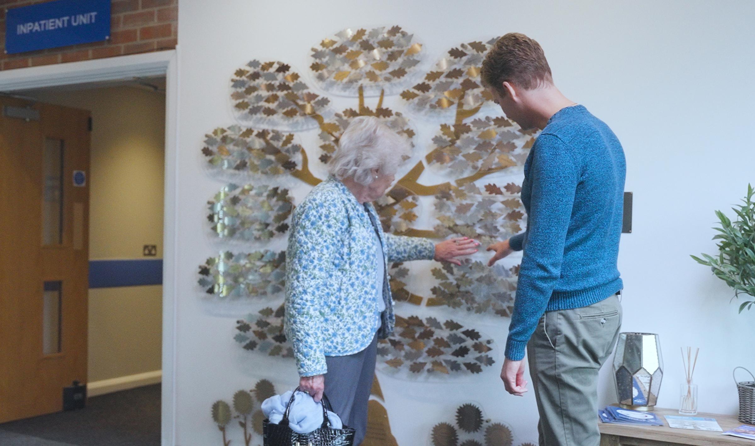 An older woman and man point at a metallic donor tree wall art in a hospital hallway.