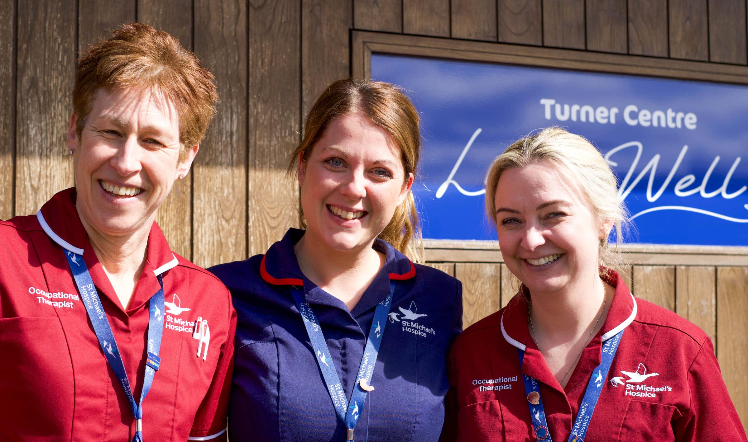 Three smiling nurses in uniforms stand in front of a wooden building.