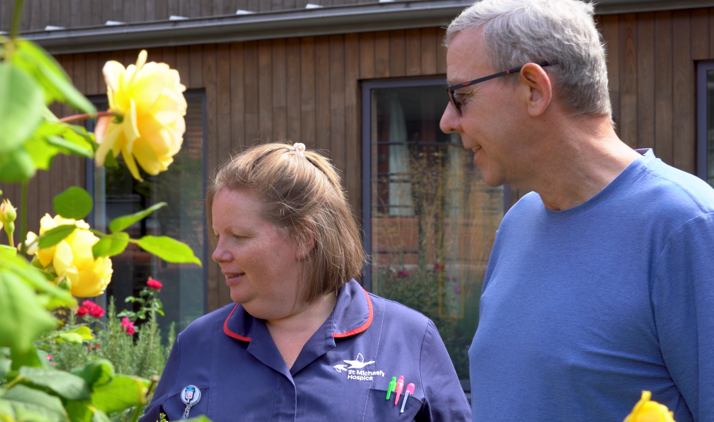 Woman in uniform and man admire yellow roses in a garden.
