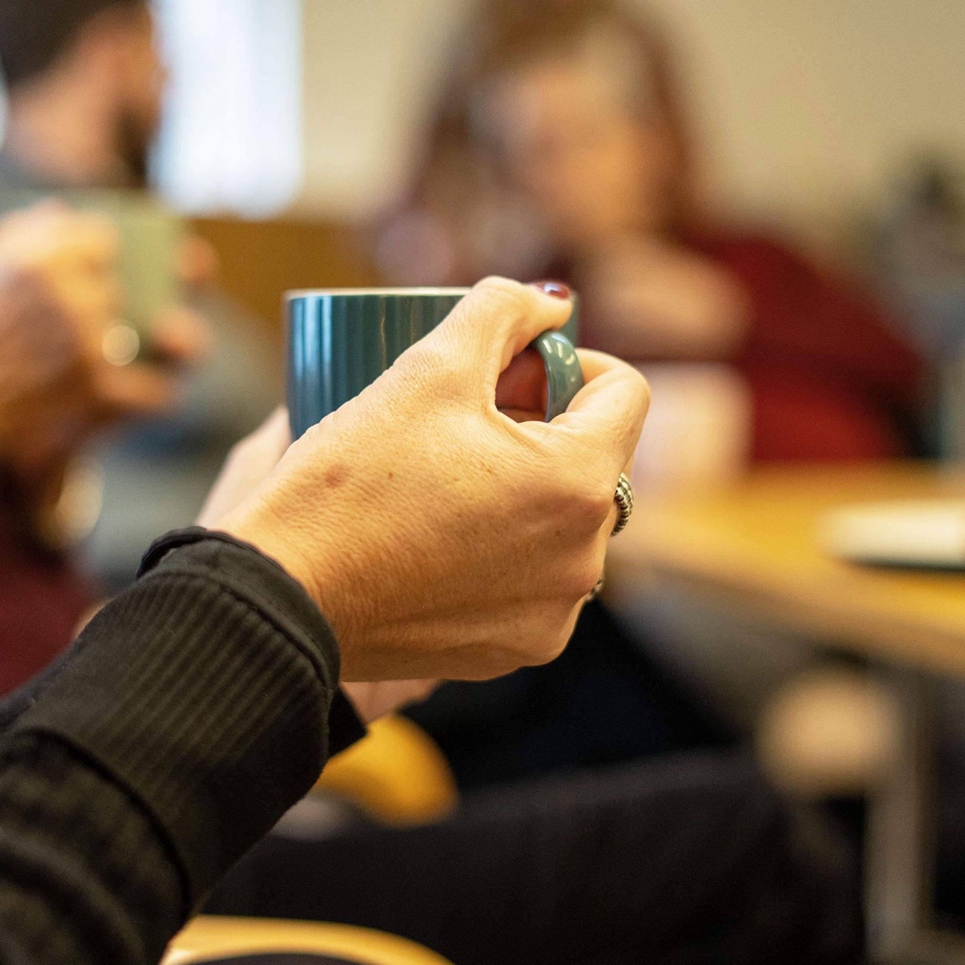 Close-up of hands holding a teal mug, blurred people in the background.