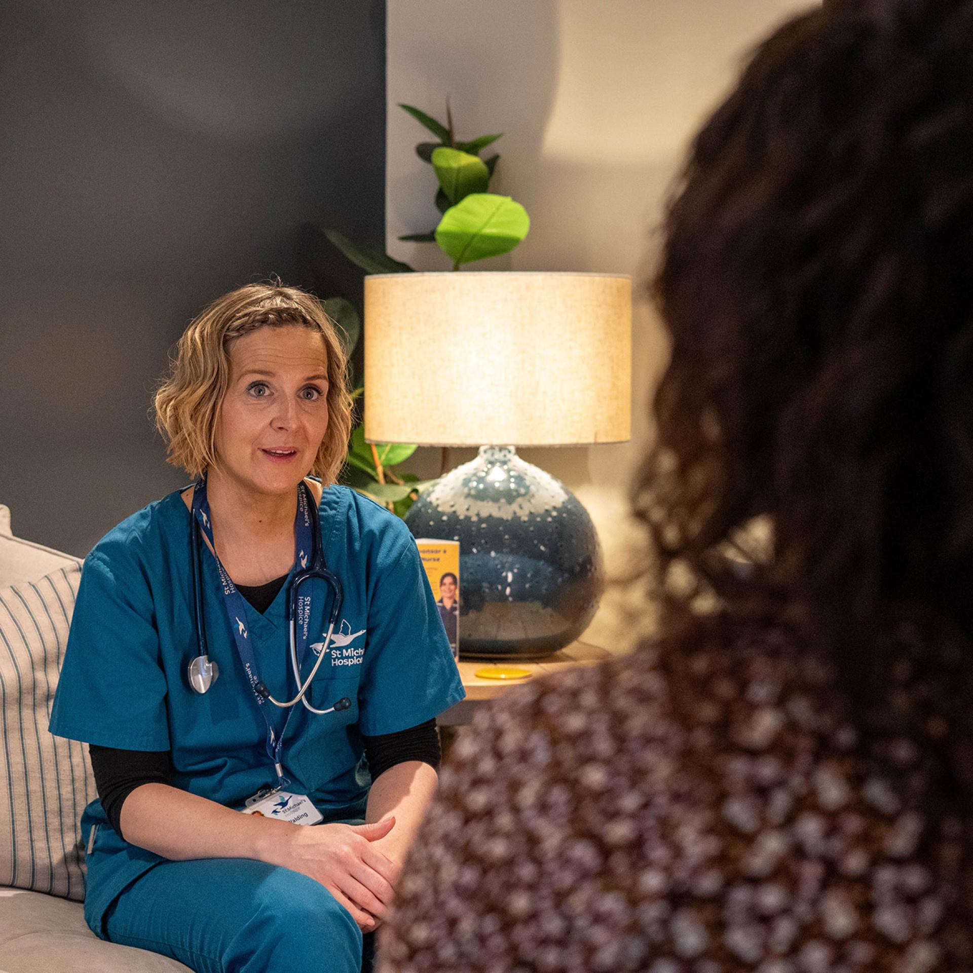 Woman in teal scrubs and stethoscope talking to another person indoors. Photograph.