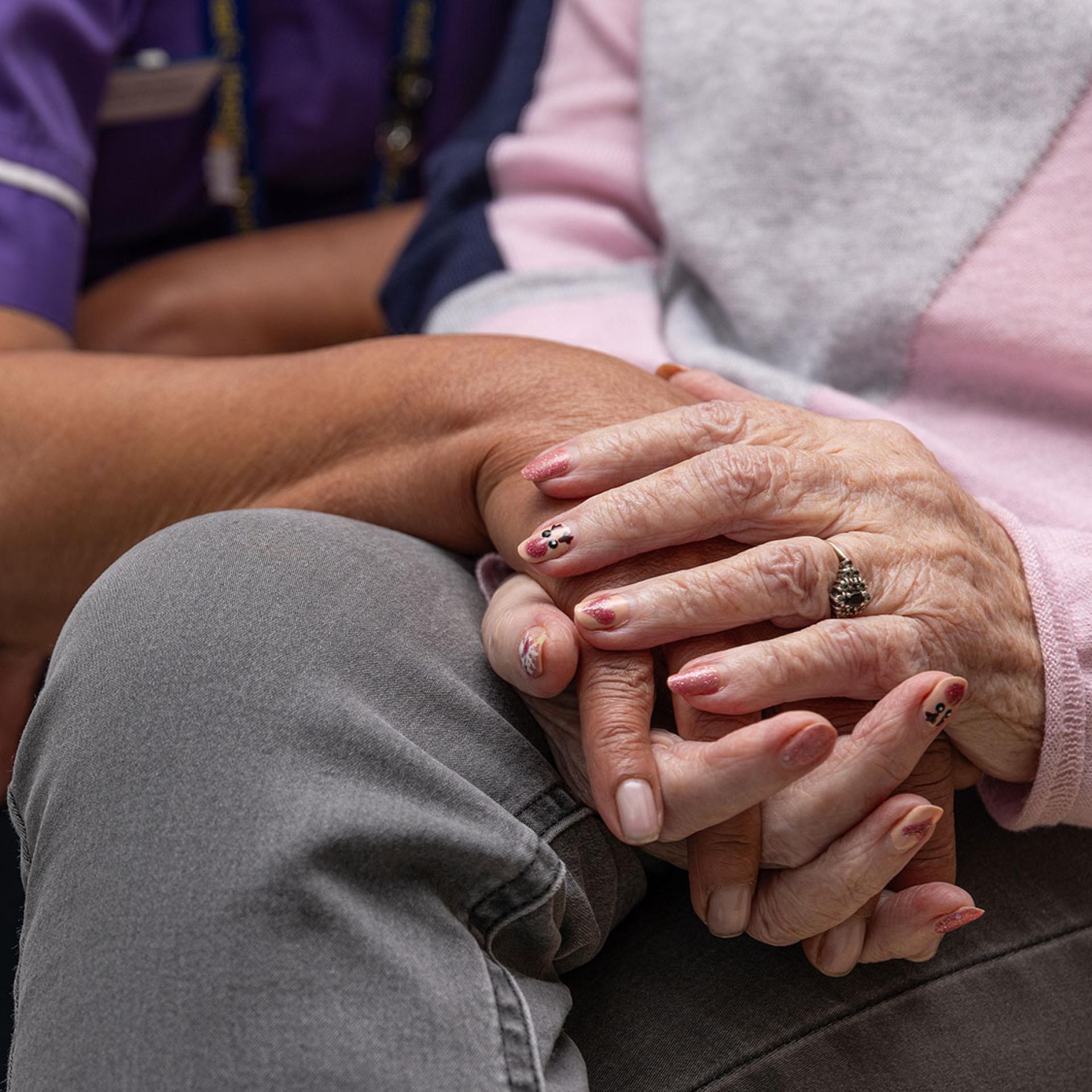 A person in a purple uniform gently holds the hand of an elderly person.