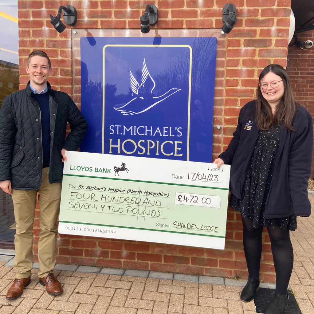Image of two people smiling at the camera with a giant check, in front of the Hospice 
