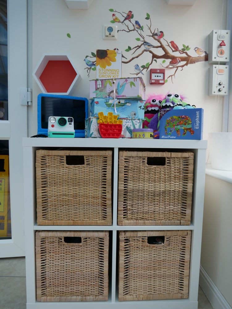 Image of the children's area in our Hospice, featuring a storage shelf with wicker boxes, on top of the unit is several resources