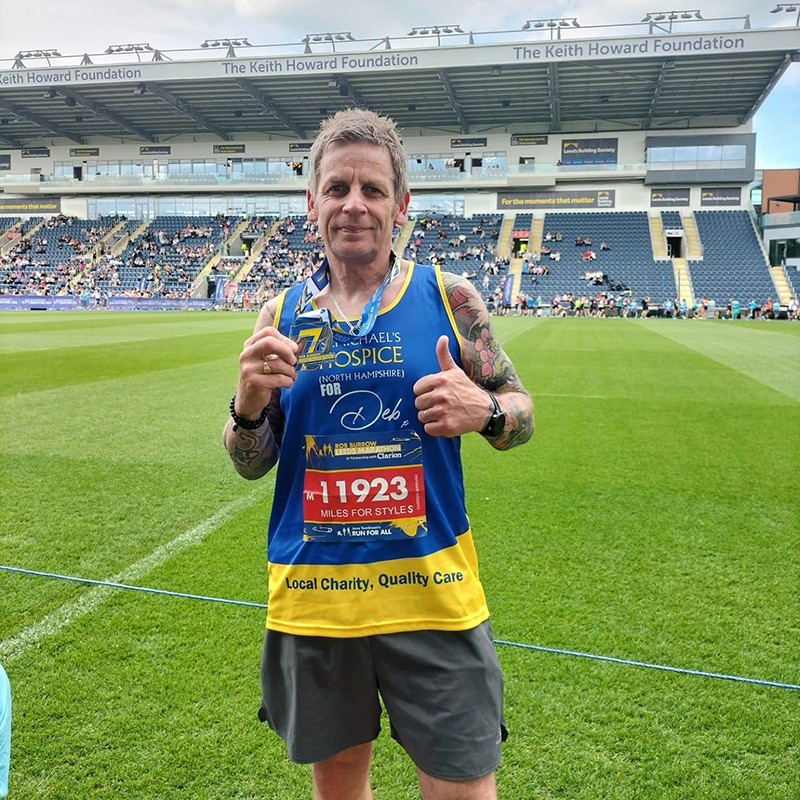 Image of Darren holding his medal and smiling at the camera. He has a thumb up and is wearing at St. Michael's Hospice running vest.