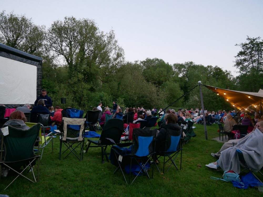 Image of people sat outside at sunset ready to watch a film on a big screen outside